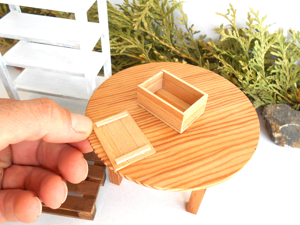 A miniature wooden table and matching box set, shown with a human hand holding one piece to demonstrate scale. The set is photographed against a white background with decorative green foliage and a miniature white shelf.