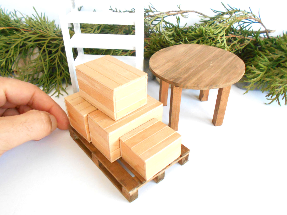 A miniature wooden toy set featuring a stack of cargo boxes on a small pallet, shown with a hand adjusting it. The set includes a small round table, a white chair, and green pine branches in the background, all on a white surface.