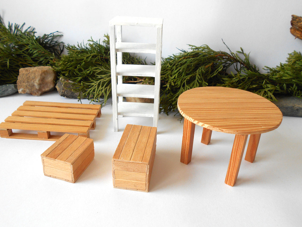 A miniature furniture set for dollhouses, featuring a round light wood table, two rectangular wooden boxes, a wooden pallet, and a white wooden ladder, arranged on a white surface with green foliage and rocks in the background.