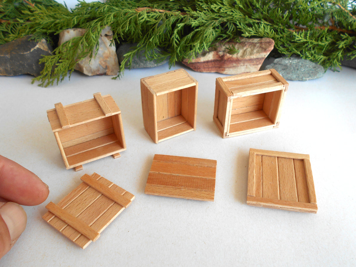 Six miniature light brown wooden crates and boxes of various sizes, including open storage crates and flat lids, arranged on a white surface with green foliage in the background. A person's fingers are visible in the bottom left corner to show scale.