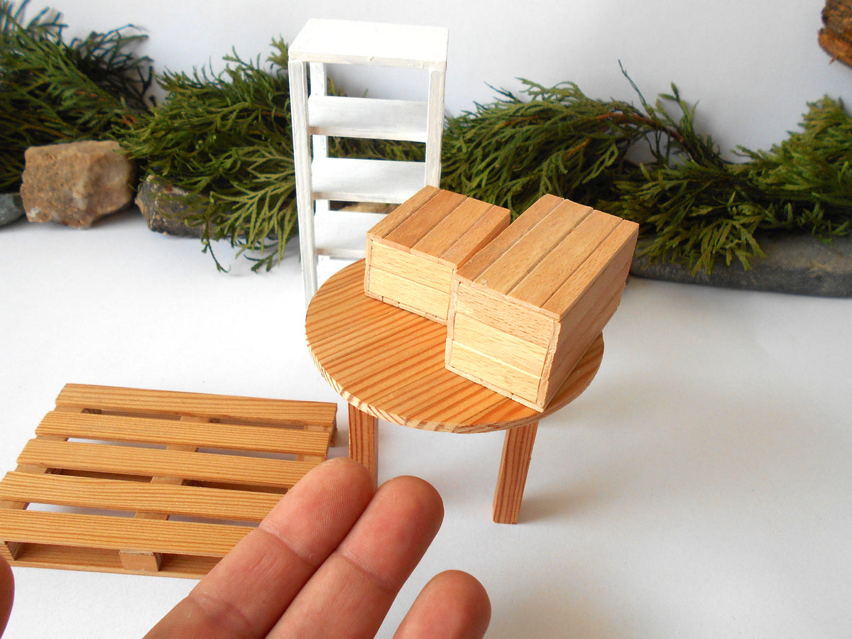 A miniature wooden dollhouse furniture set featuring a round table with two small wooden blocks, a matching pallet-style tray, and a white ladder, shown with a human hand for scale against a white background with green pine garland.