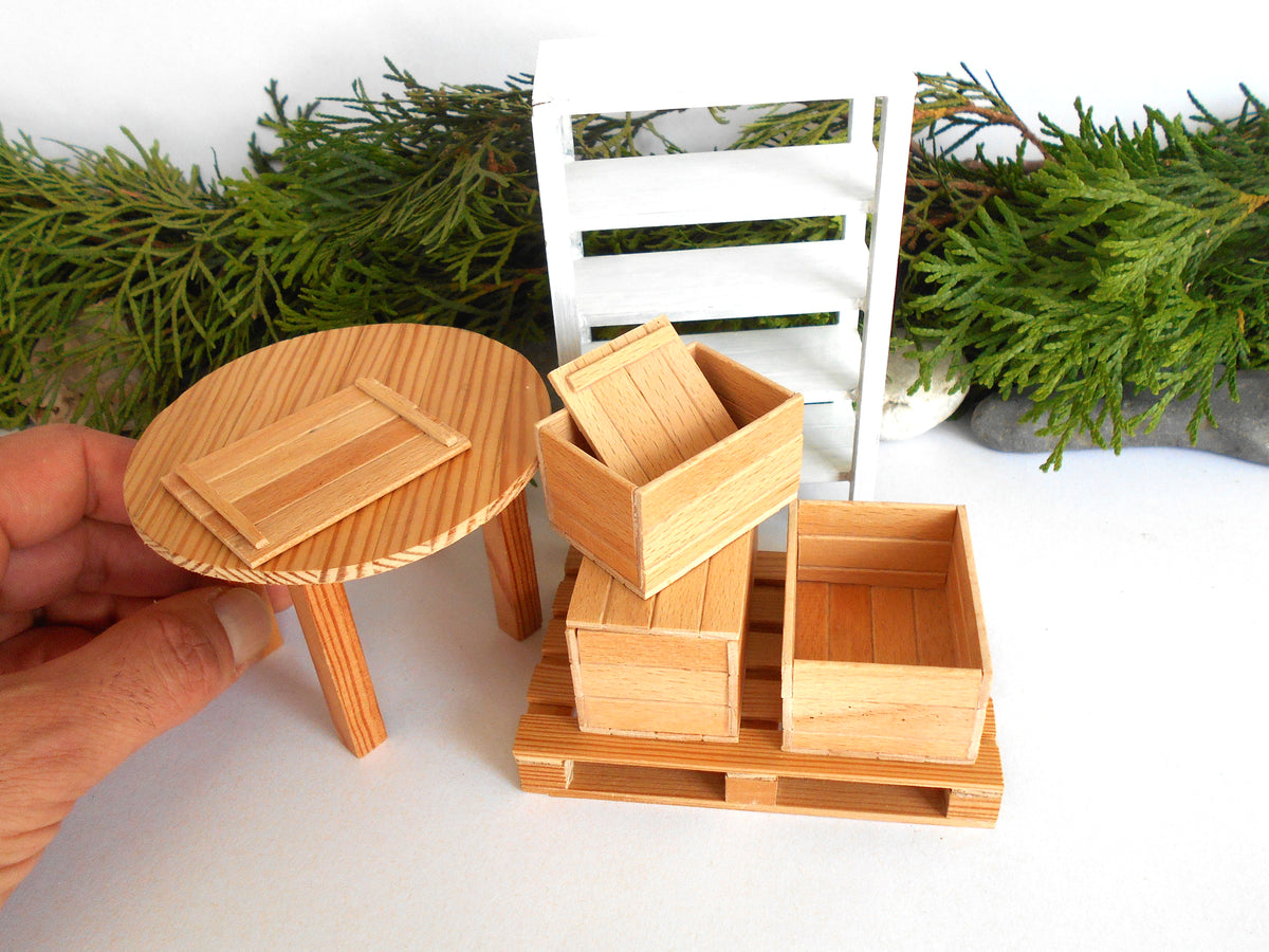 A miniature wooden dollhouse furniture set including a round table, a stack of storage crates, and a pallet, shown with a hand holding part of the table and decorative greenery in the background.