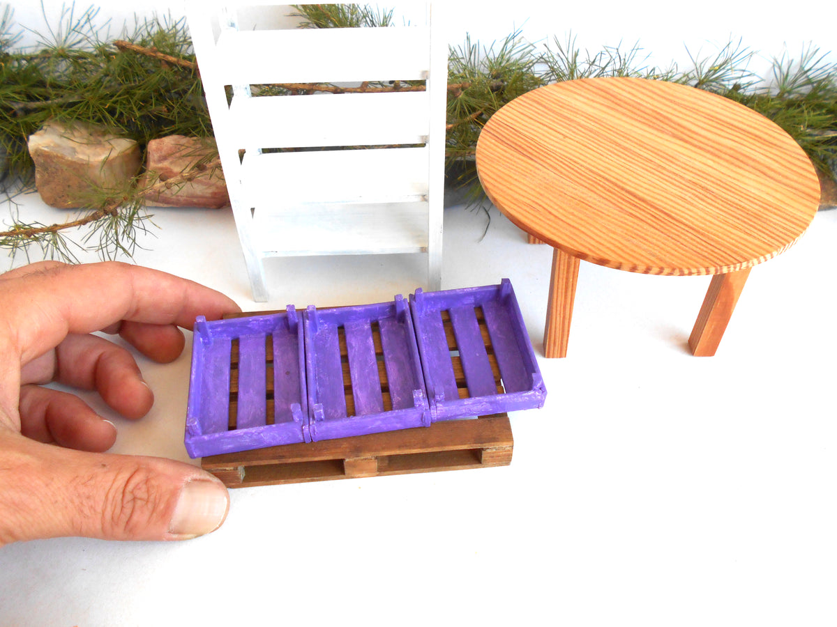 A miniature purple slatted bench or crate-like toy piece resting on a small wooden base, held by a human hand in the foreground. A white chair, a round wooden table, and pine branches are visible in the background against a white surface.
