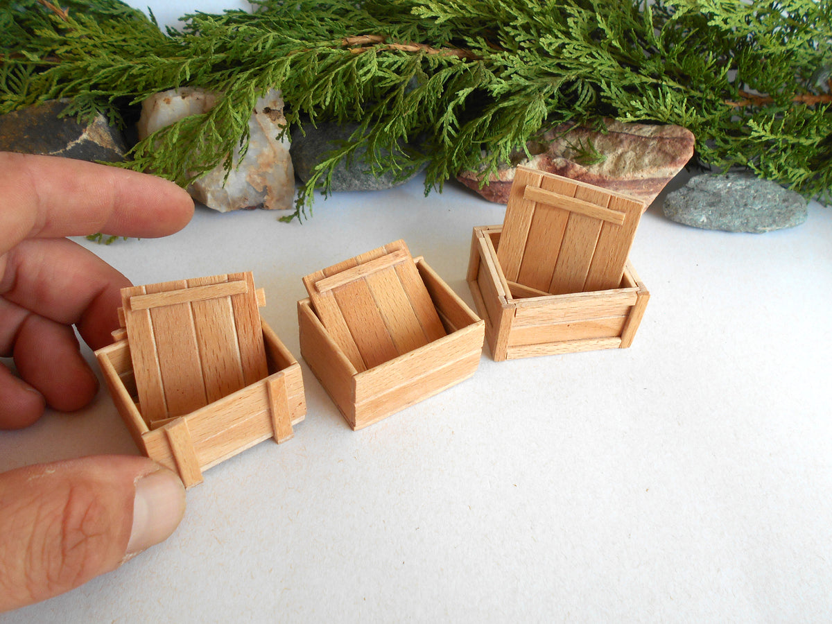 Three miniature light wood crates or boxes displayed on a white surface, with a hand holding one on the left for scale and green foliage in the background.