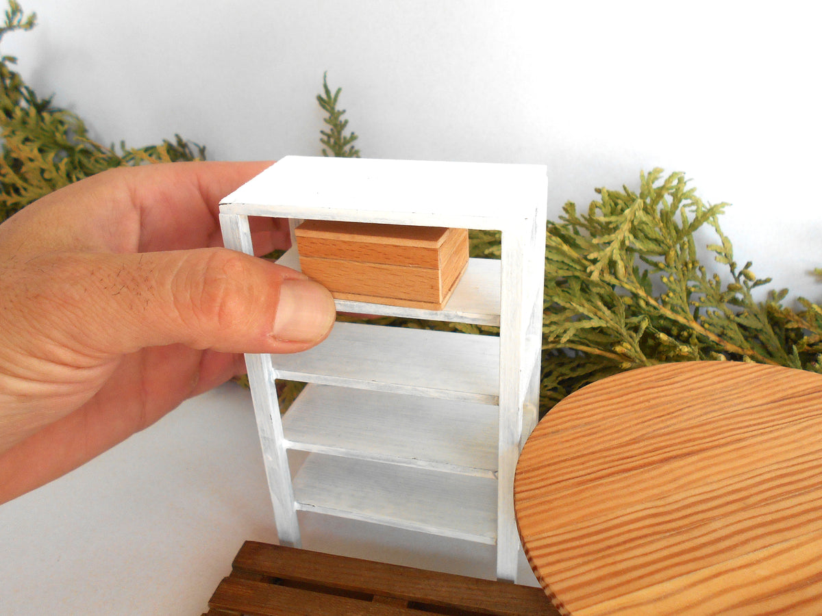 A miniature white three-shelf bookcase with a small wooden box on the top shelf, held by a hand to show scale, with a wooden table and green foliage in the background.
