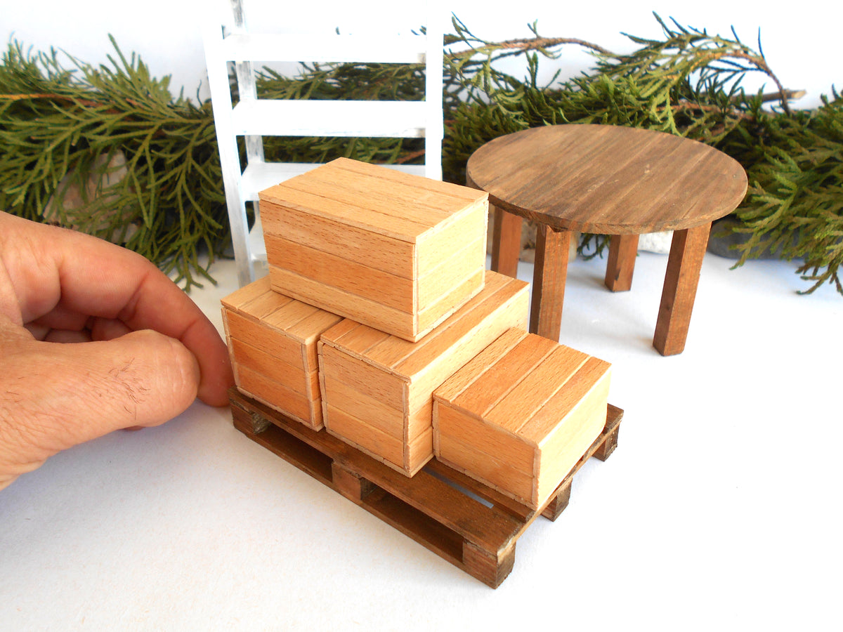 A miniature wooden toy set featuring a stack of three small cargo boxes on a pallet, with a hand holding it for scale, alongside a miniature round table and chair against a white background with green pine branches.