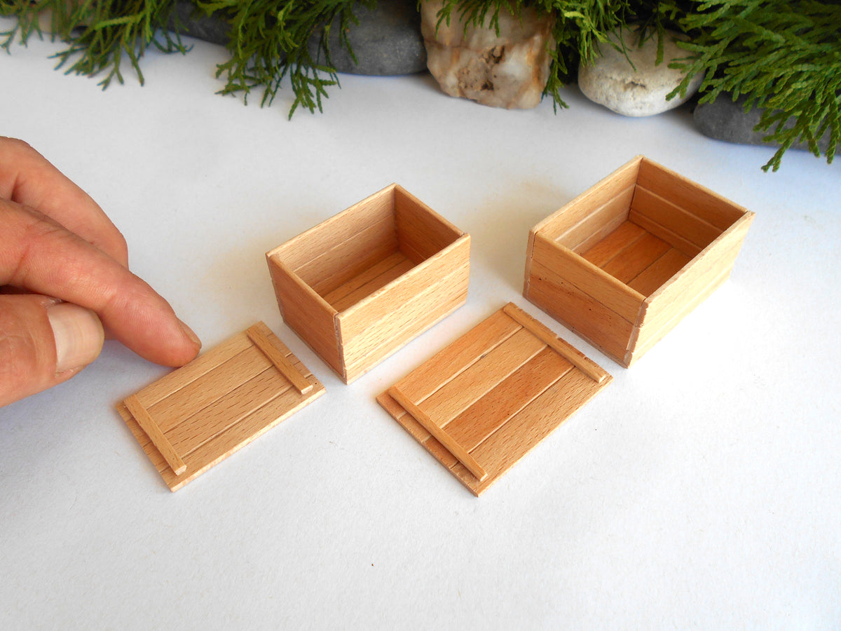 Four miniature light wood boxes and lids, including one shown being held by a hand to demonstrate scale, arranged on a white surface with decorative greenery in the background.