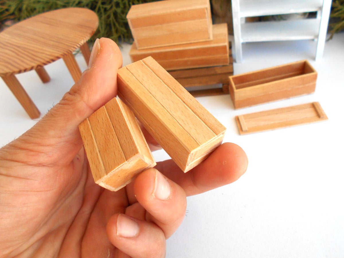 A hand holding two small wooden miniature furniture pieces, with other matching wooden toys and a white shelving unit blurred in the background.