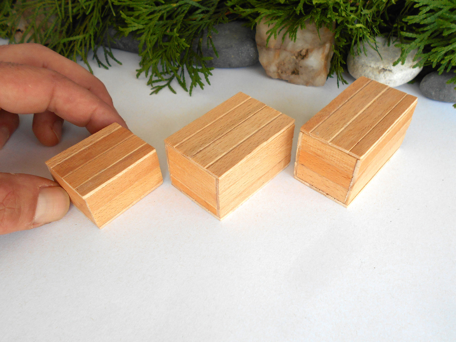 Three small rectangular wooden boxes with a striped pattern, displayed on a white surface with a hand holding the smallest box. Green foliage and stones are visible in the background.