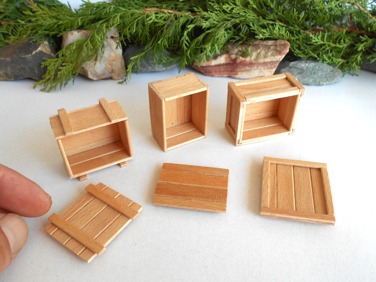 Six miniature wooden crate and door pieces arranged on a white surface, with a hand holding one piece at the lower left and evergreen branches and stones in the background.