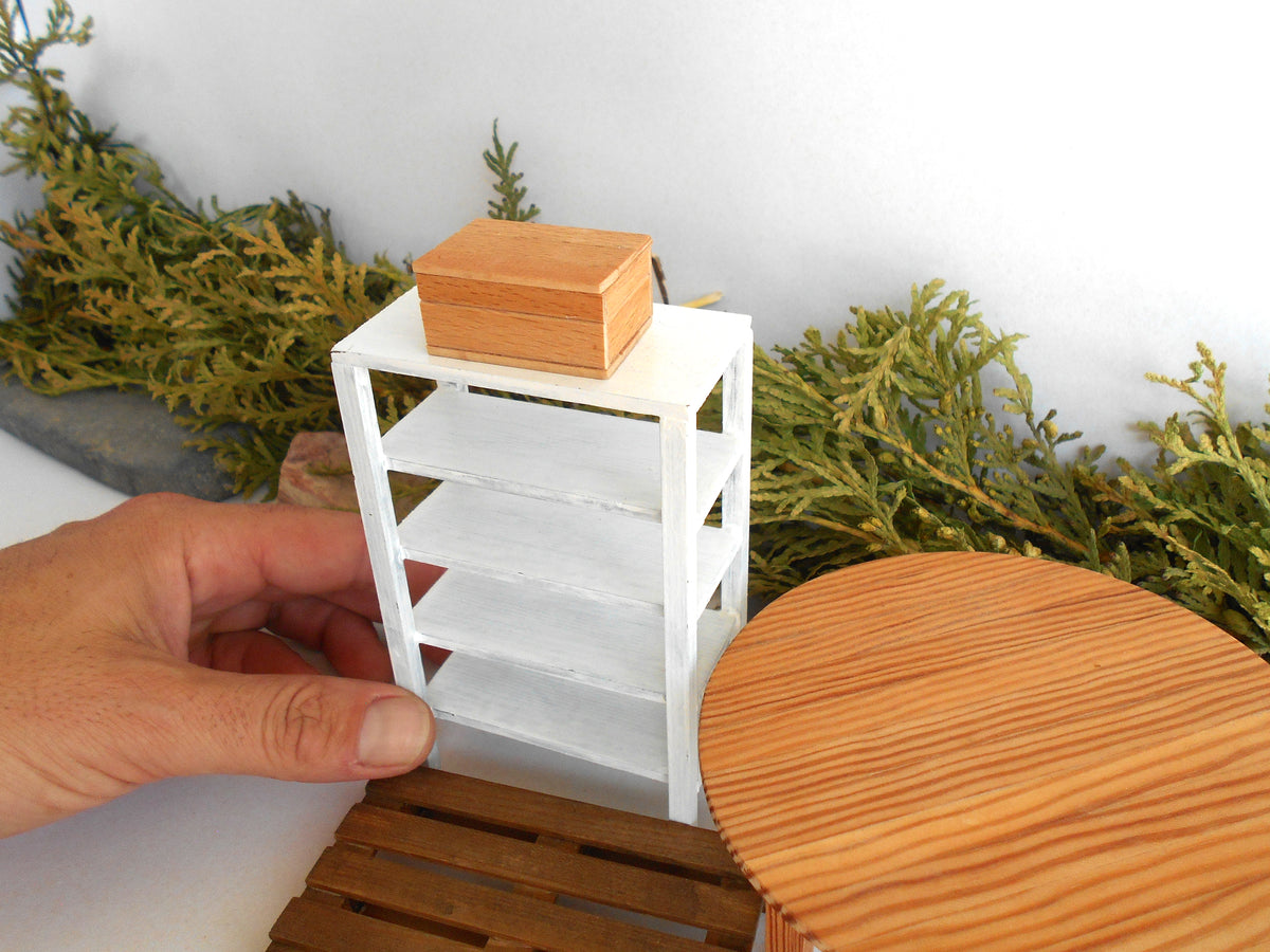 A miniature white shelving unit with a small wooden box on top, held by a hand in a miniature diorama setup with faux greenery and a wooden table.