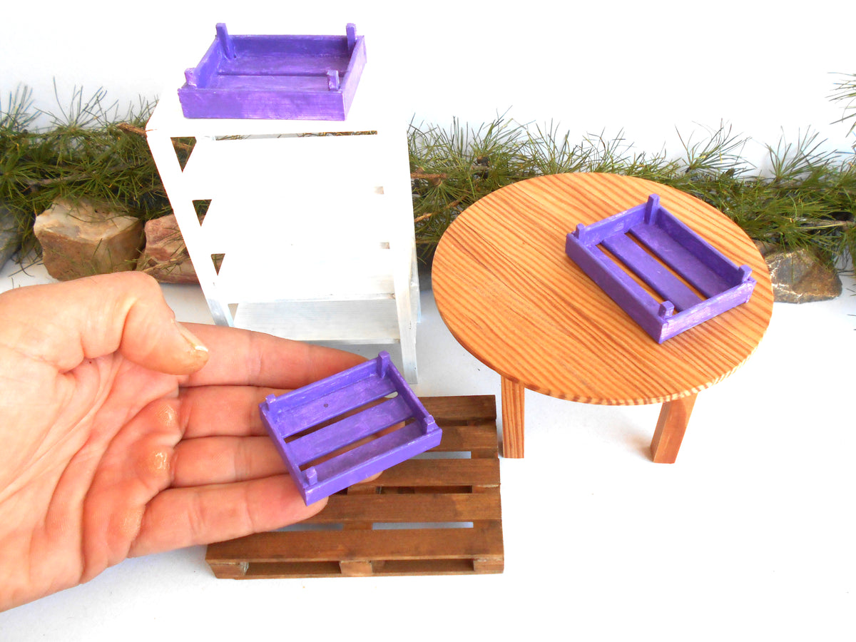 A miniature purple crate toy shown in a hand, with two more miniature purple crates placed on a small wooden table and a miniature white shelf, against a white background with decorative greenery.