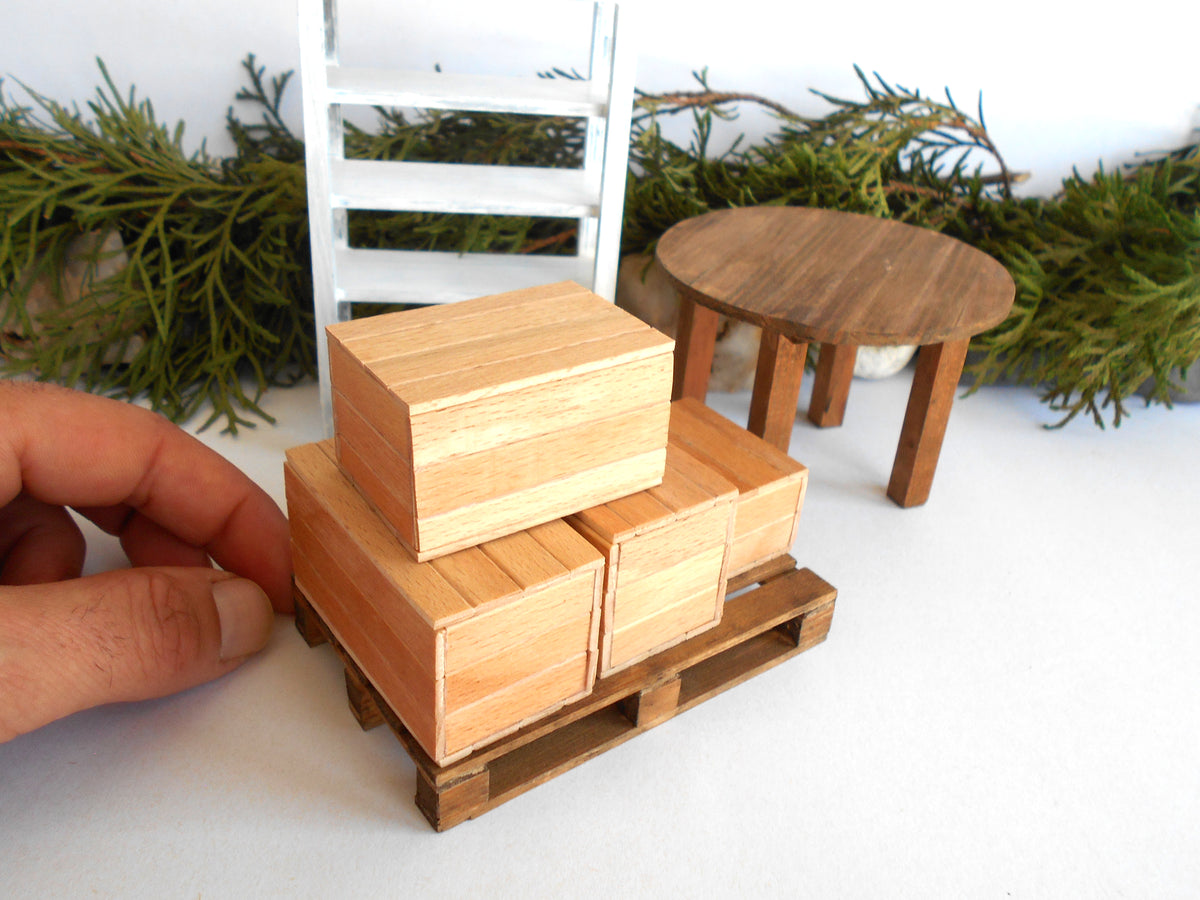 A miniature wooden toy building set featuring stacked boxes on a pallet, shown with a hand for scale next to a small round table and ladder.