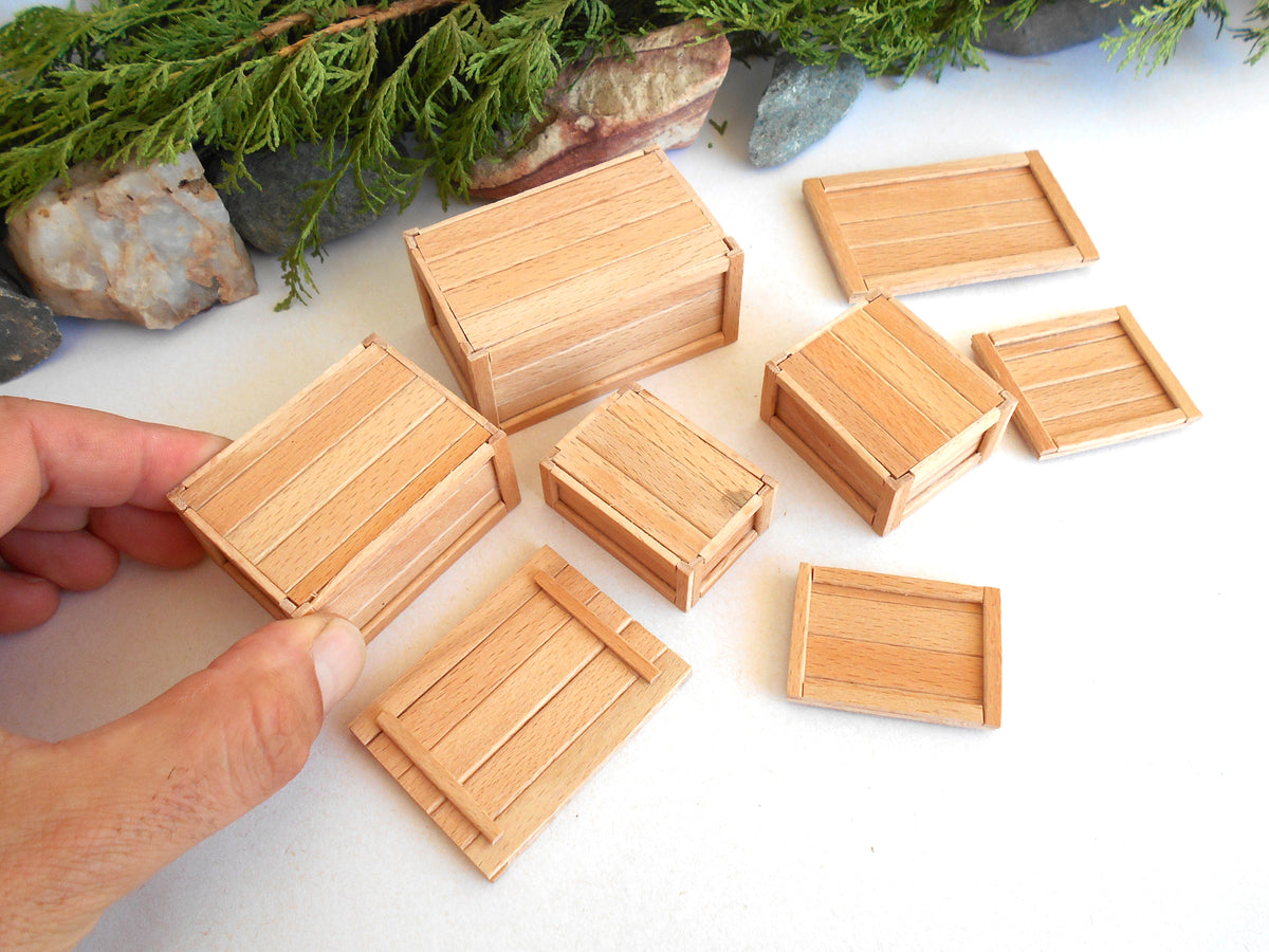 A hand holding a miniature light wood crate, with several other matching small wooden crates scattered on a white surface. Green pine branches and stones decorate the top of the scene.