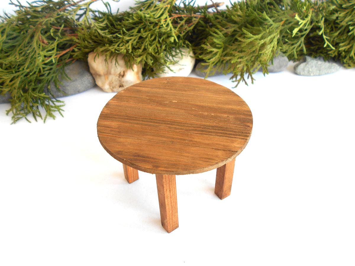 A miniature round wooden table with three legs, styled on a white surface with green foliage and small stones in the background.