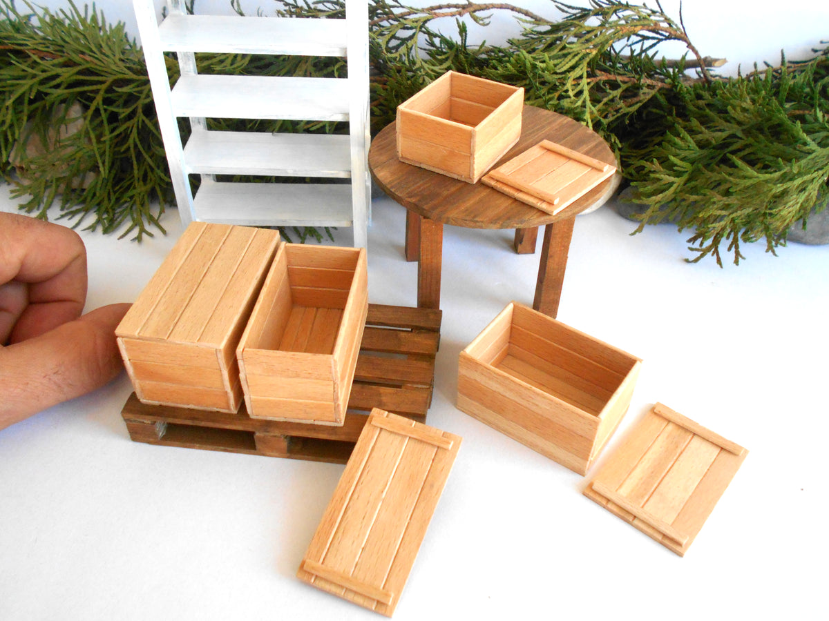 A set of miniature wooden crates and boxes with matching lids, displayed on a white surface with a small white chair, a round wooden table, and green foliage in the background. A human hand is visible on the left side of the frame.