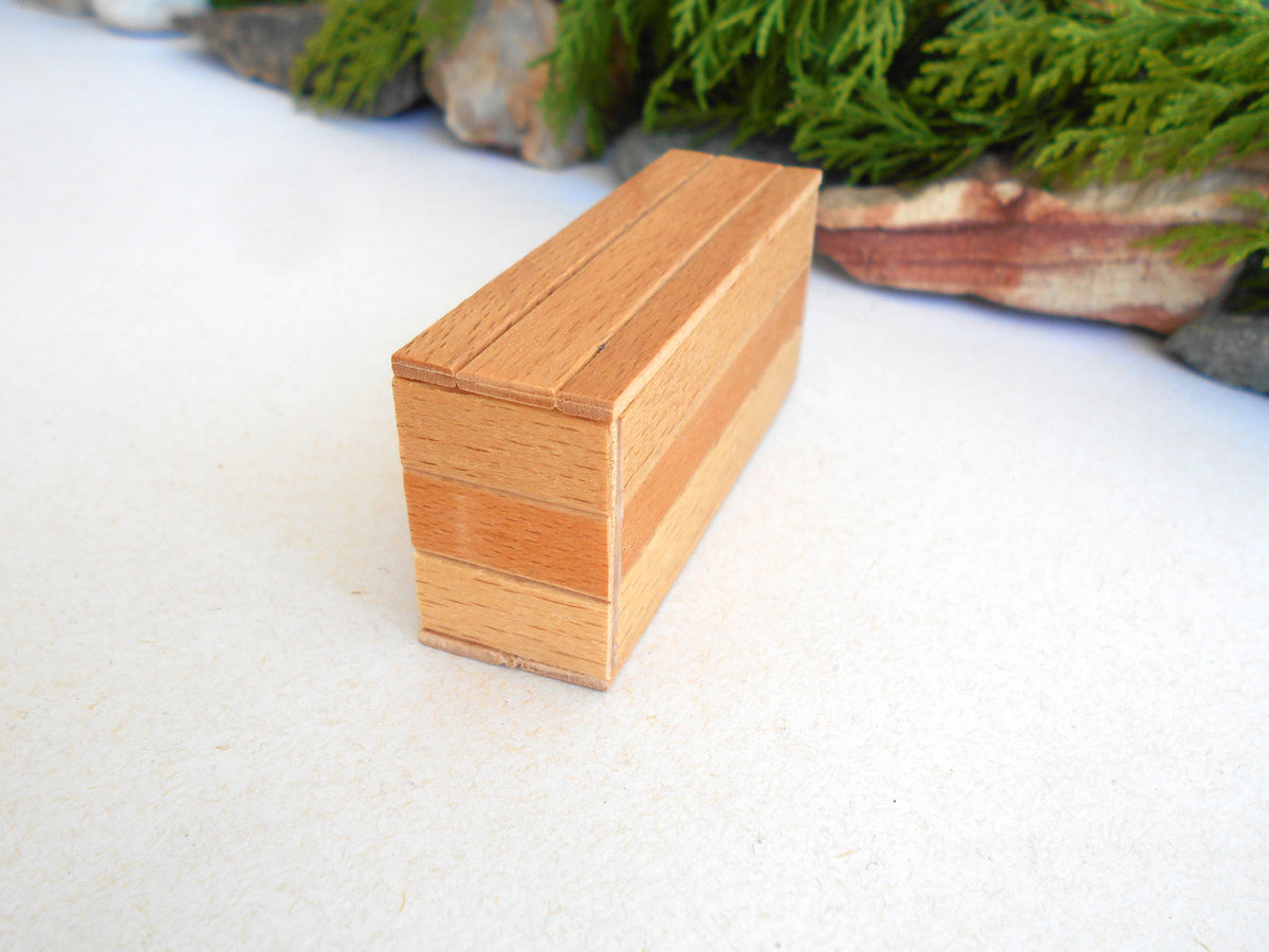 A small rectangular wooden box with a herringbone pattern, standing on a white textured surface with blurred green foliage in the background.