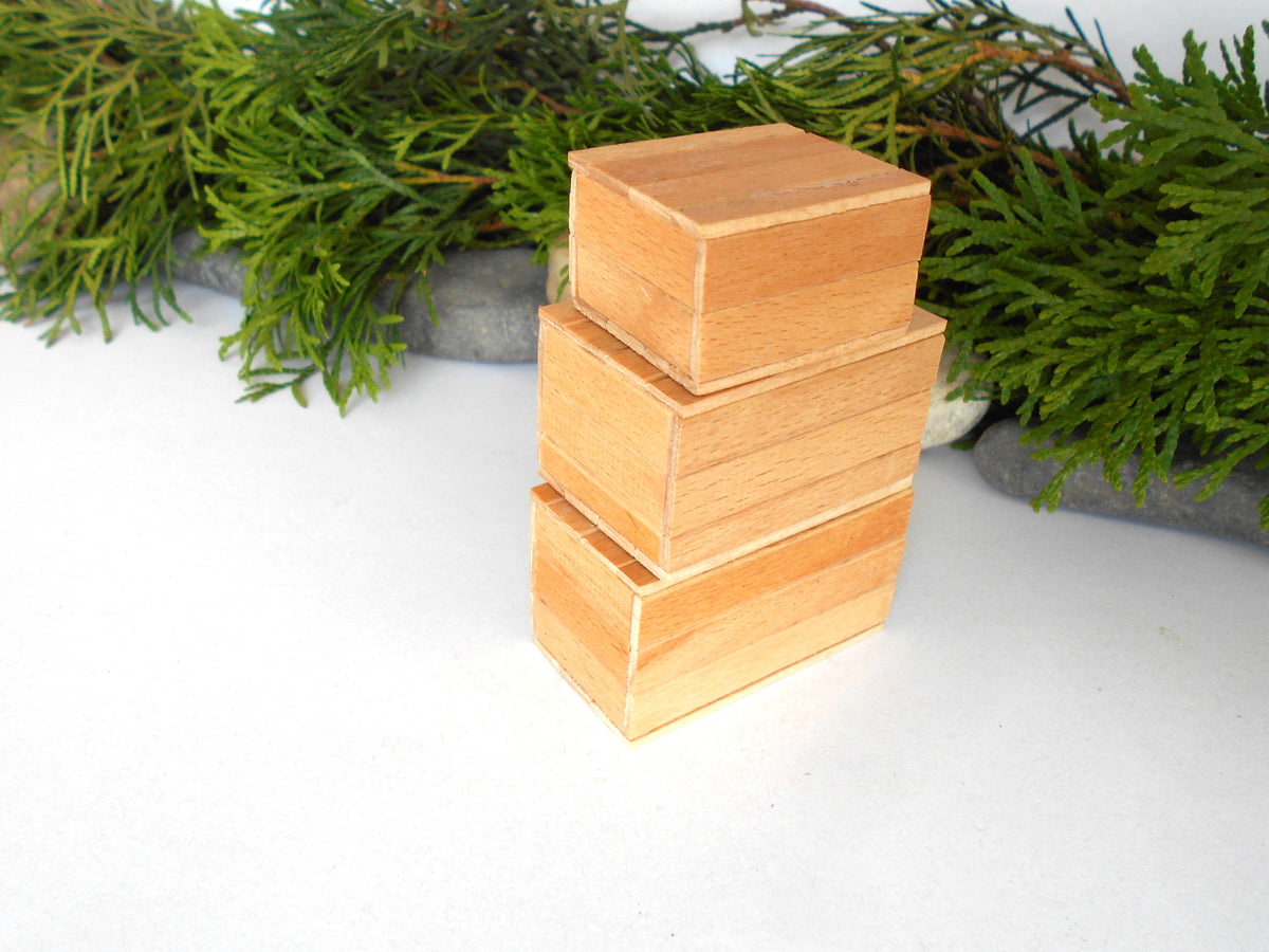 Three small stacked wooden boxes in a warm light tan color, set against a white background with green pine branches in the background.