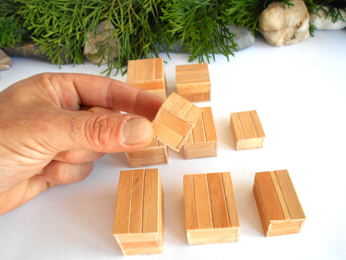 A hand holding a small wooden boxes , with several matching miniature wooden boxes arranged on a white surface in front of green foliage and stones.