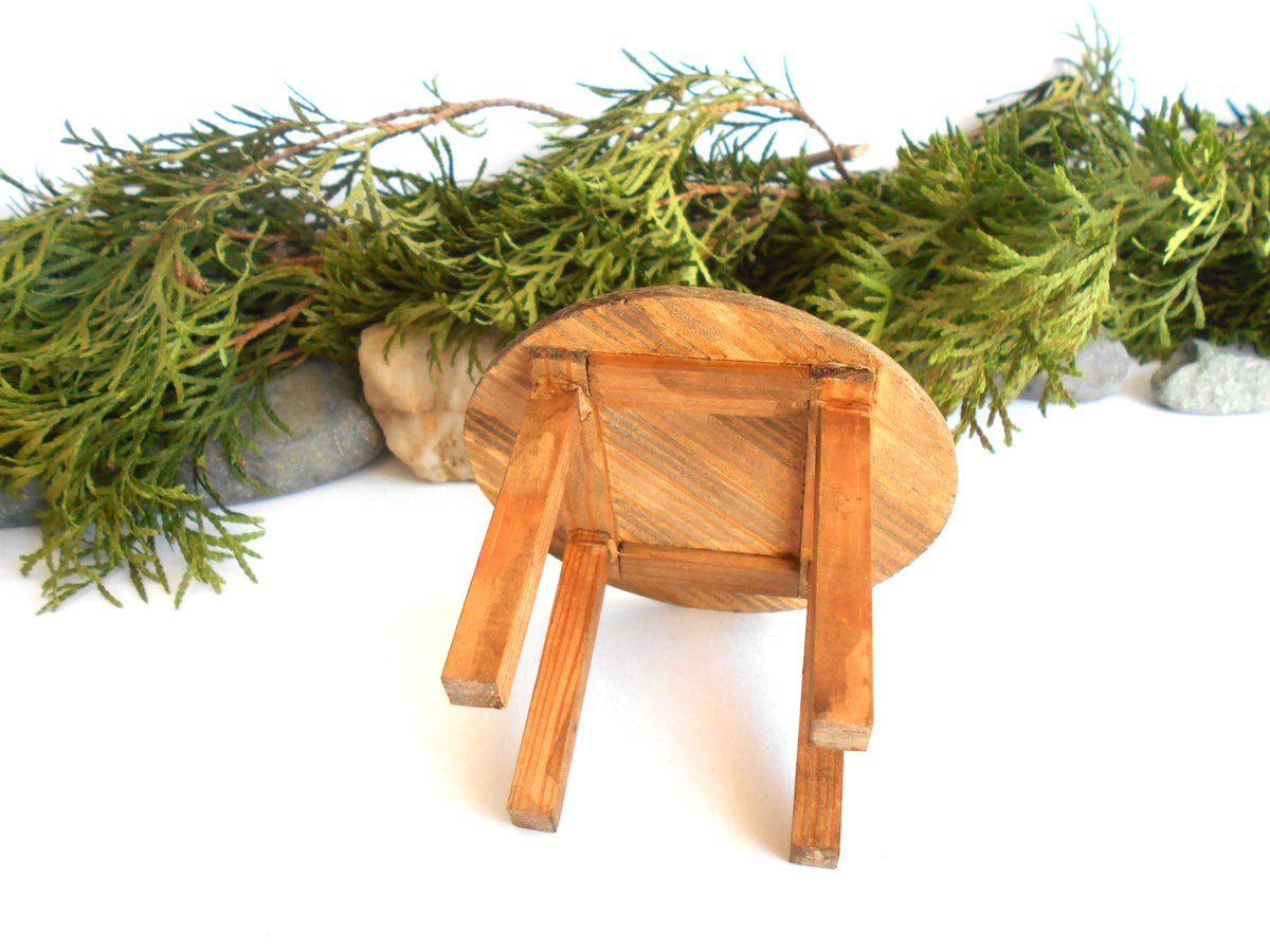 A miniature wooden stool with a round seat and four legs, shown from a low angle against a white background with green foliage and small stones as decorative props.