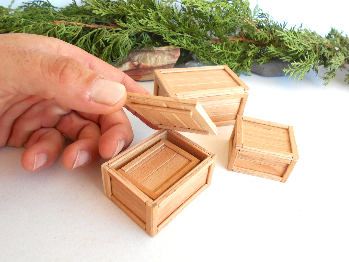A close-up of a hand holding open one of several small wooden crates on a white surface, with green foliage in the background.