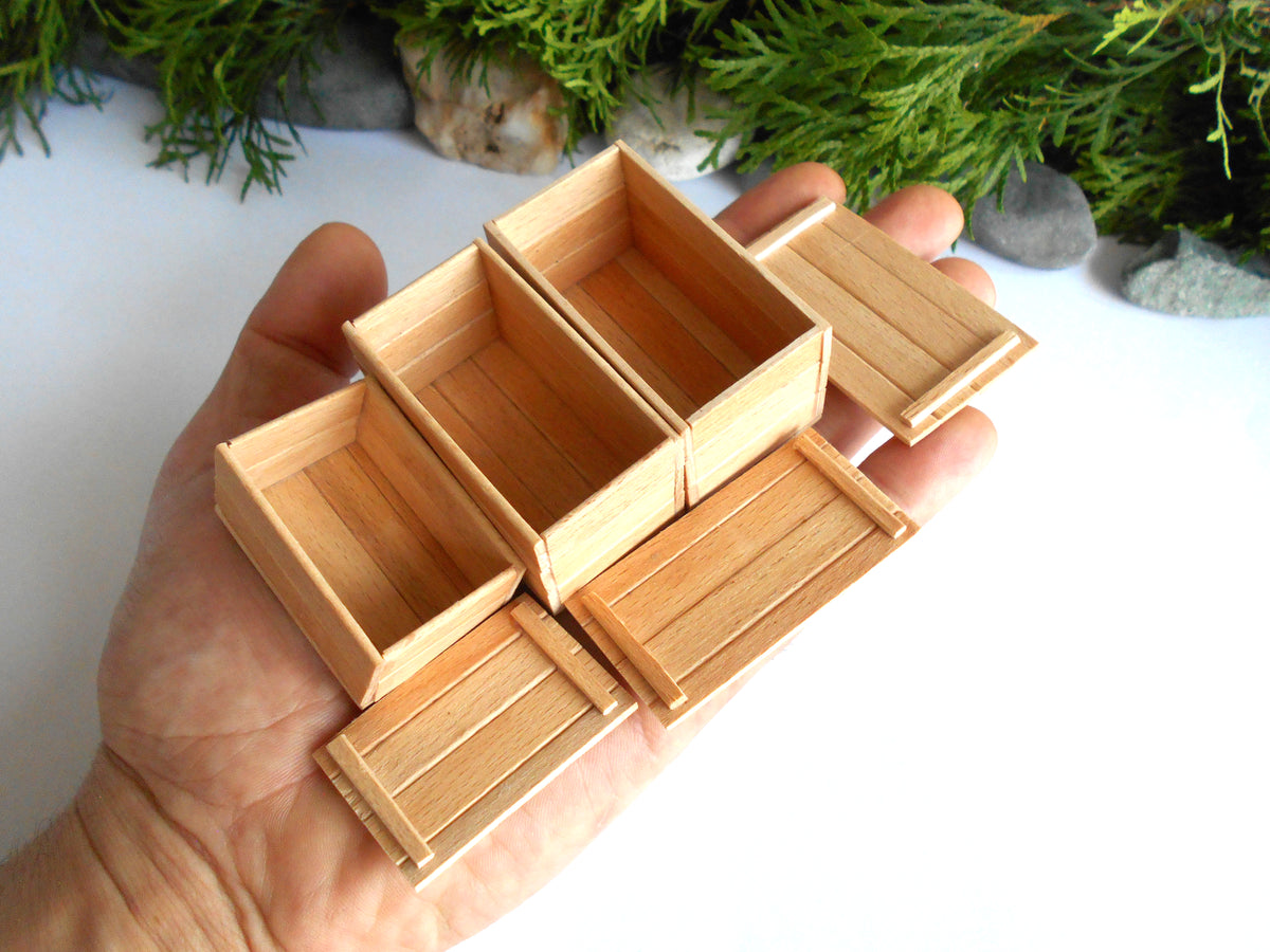 A hand holding a set of miniature light wood boxes and trays, with green foliage and small stones in the background.