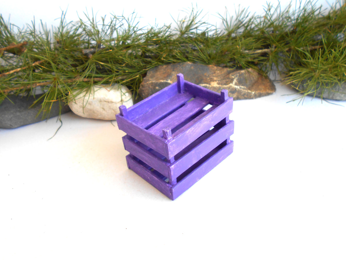A small purple wooden slat crate or basket shown on a white surface with decorative green pine branches and stones in the background.