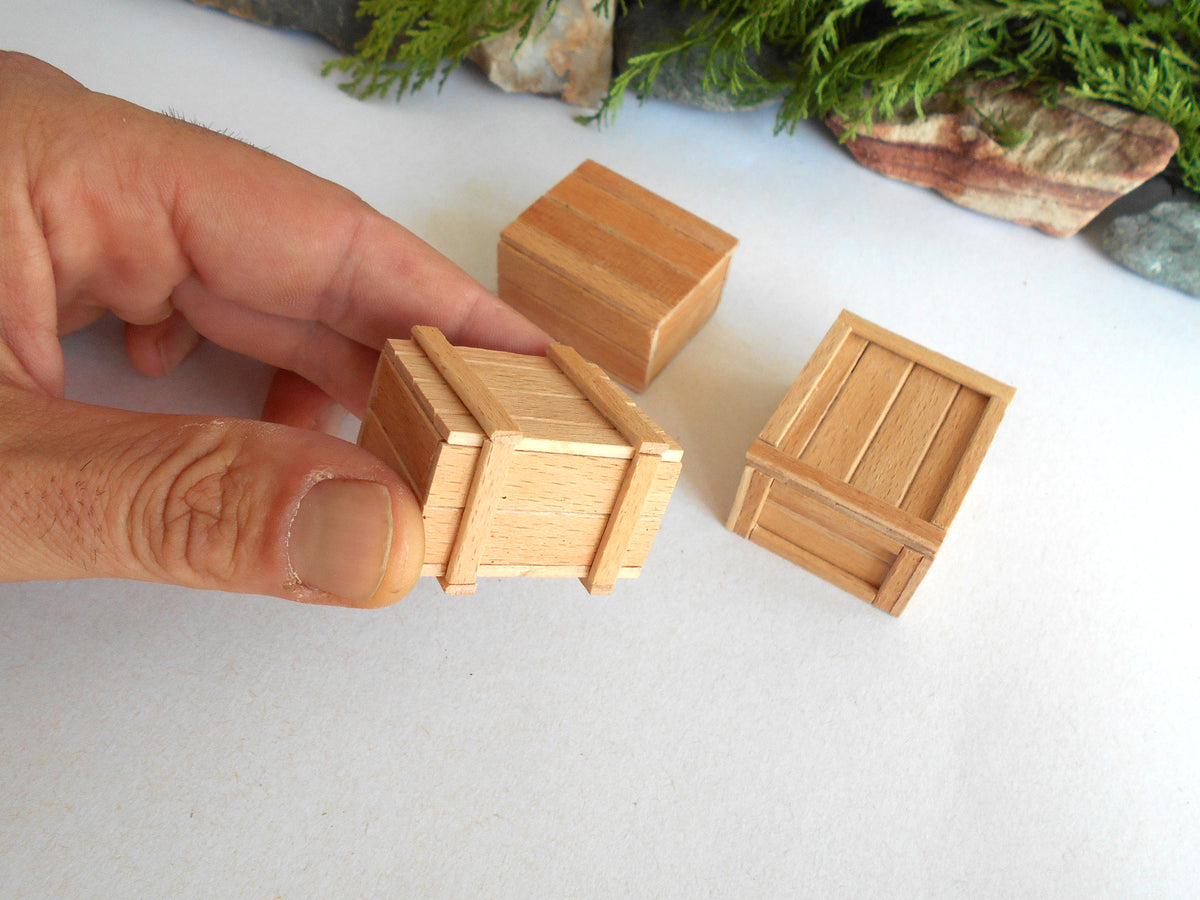 A close-up of a hand holding a miniature wooden crate, with two more matching small wooden crates on a white surface in the background.