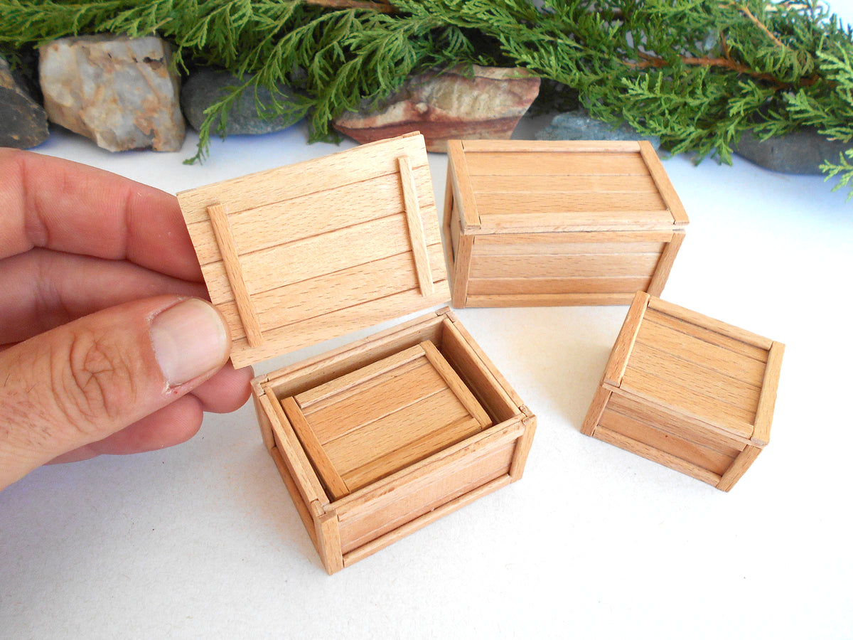 A hand holds an open miniature wooden crate box next to three closed matching crate boxes on a white surface, with green foliage and small rocks in the background.
