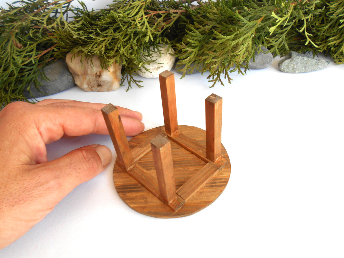 A miniature wooden stool or small table stand with a round base and four legs, shown on a white surface with a hand holding it for scale and greenery in the background.