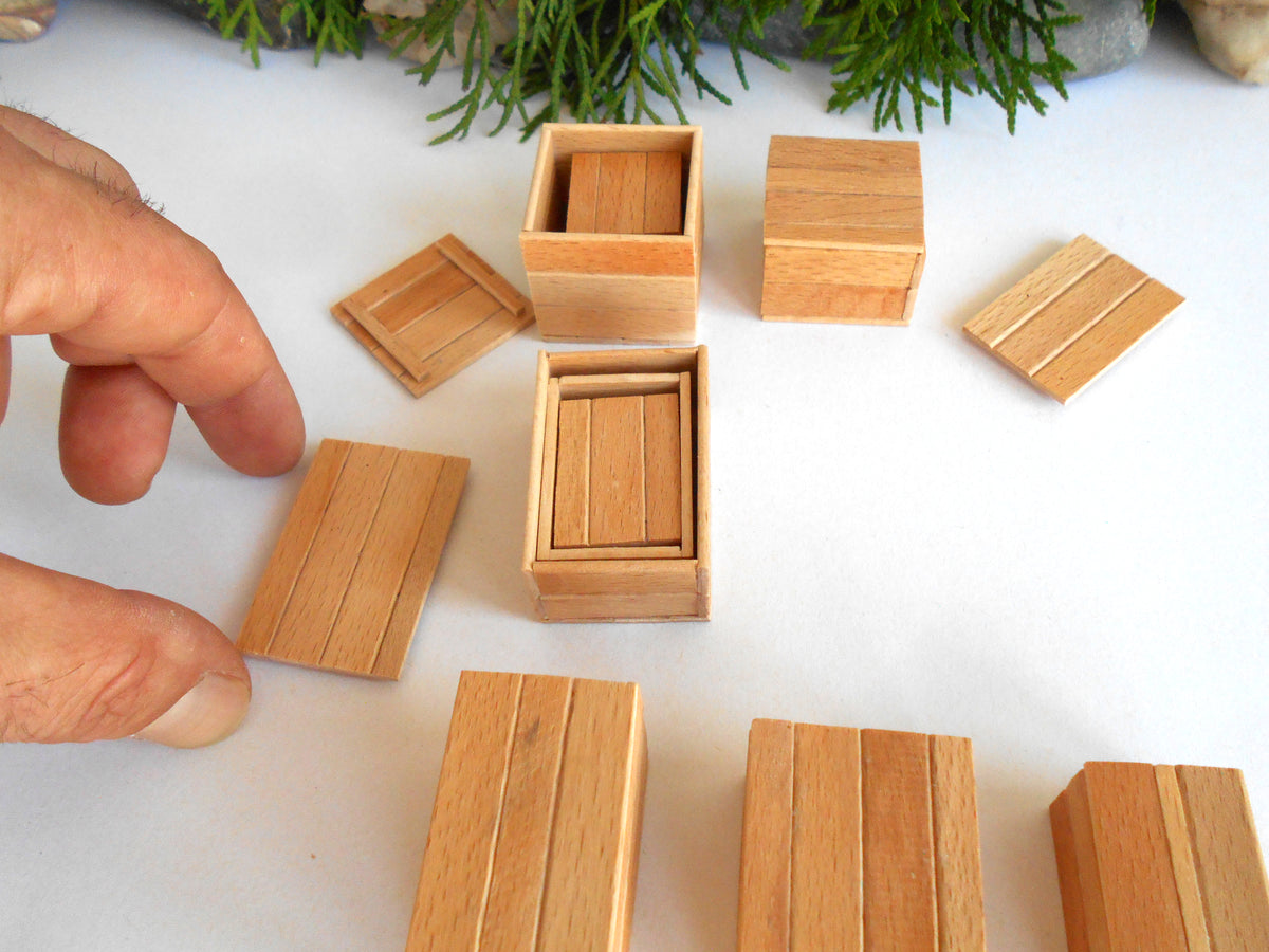A close-up of a hand arranging small light wood boxes and boxes on a white surface, with greenery visible at the top edge.