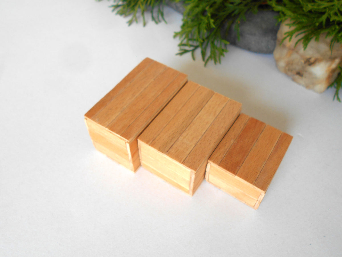 Three small rectangular wooden boxes with visible wood grain, arranged side by side on a white surface with blurred greenery in the background.