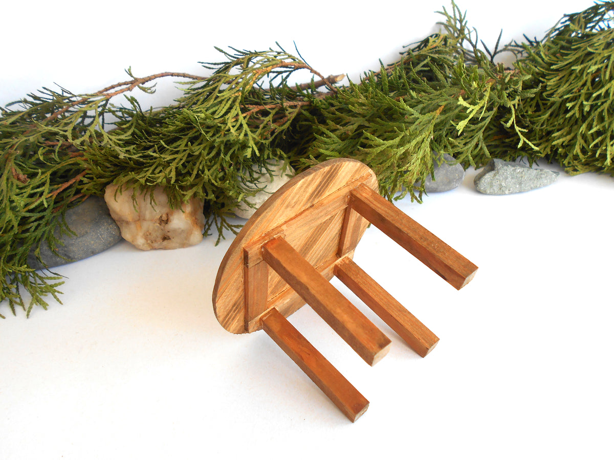 A small wooden stool with a round backrest and four legs, photographed on a white surface with green foliage and small stones behind it.