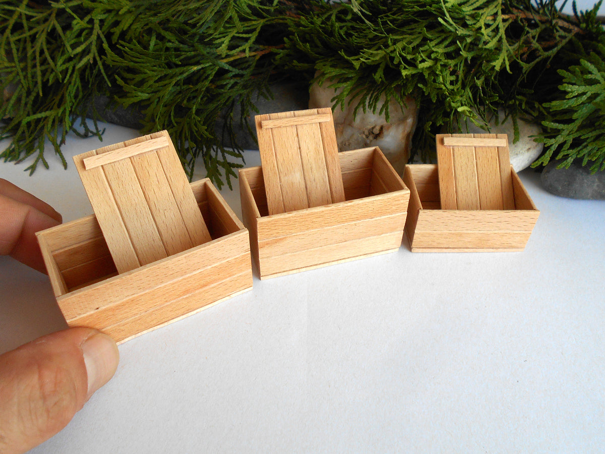 Three miniature light wood mini boxes with hinged lids, shown on a white surface with green foliage in the background. A human hand is visible holding the leftmost matchbox.