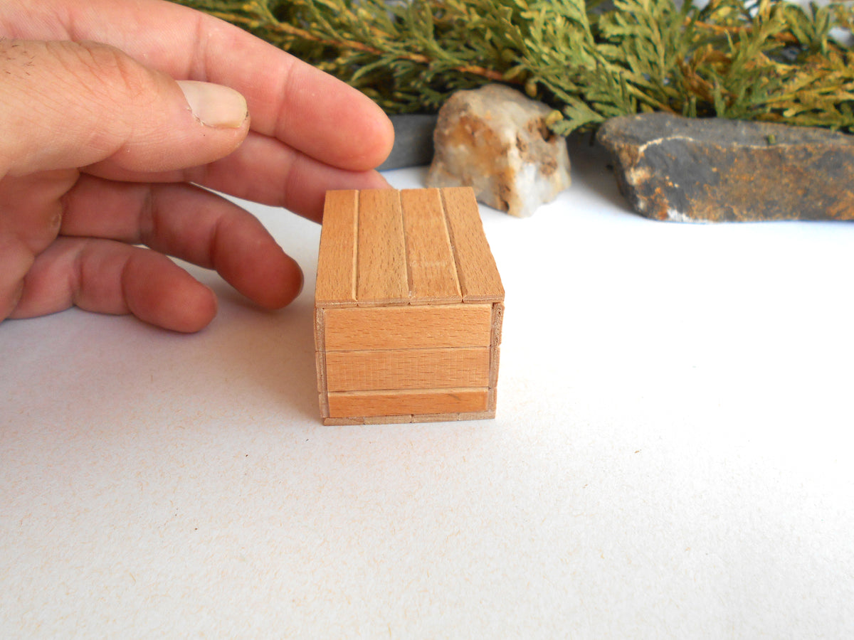 A miniature wooden box with a slatted top, held by a person's hand to show its small scale, set against a white surface with decorative stones and greenery in the background.