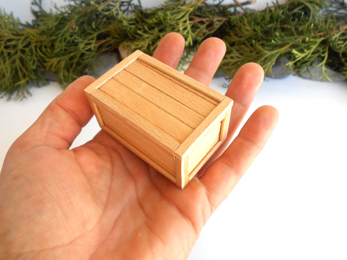 A miniature light wood box with a slatted lid, held in the palm of a hand to show its small scale, with green foliage blurred in the background.