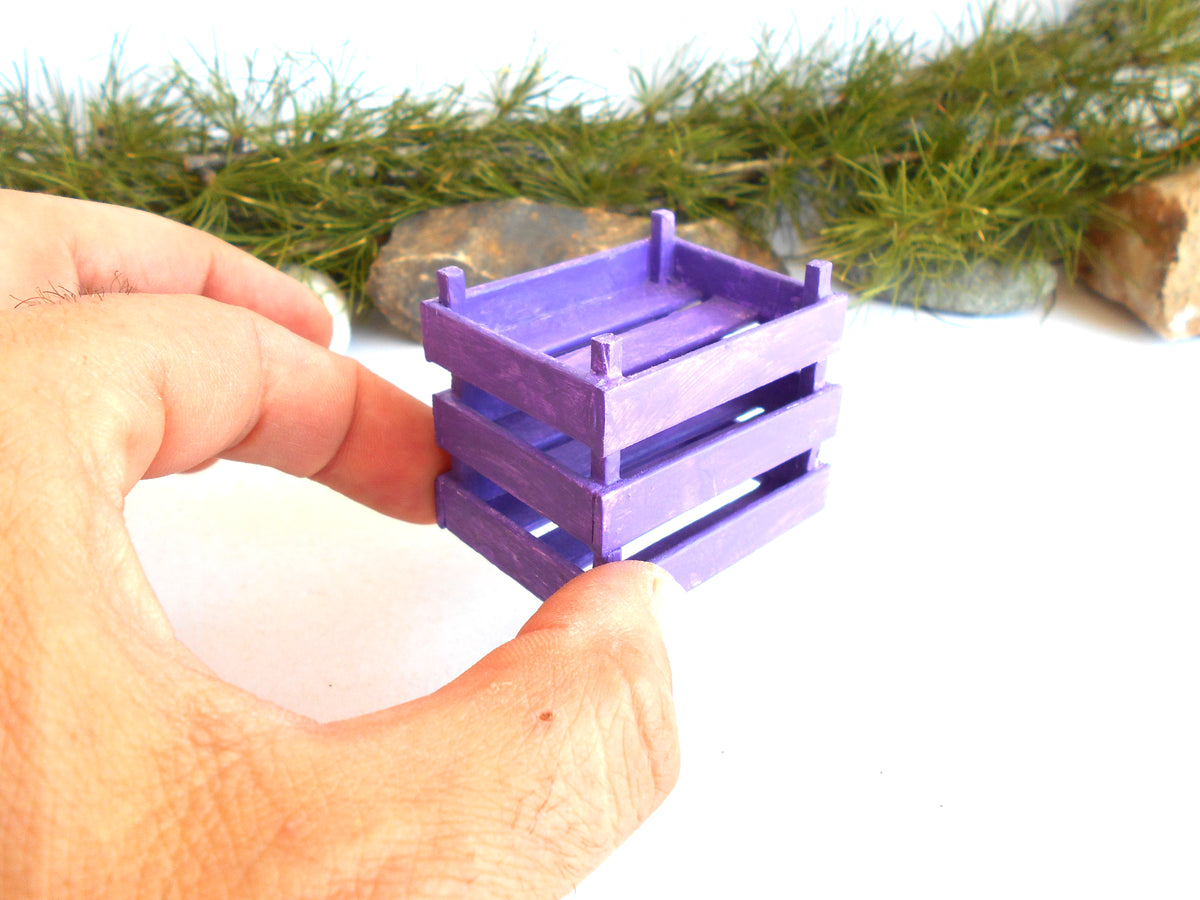 A hand holding a small purple wooden crate against a white background with green pine branches and rocks in the blurred background.
