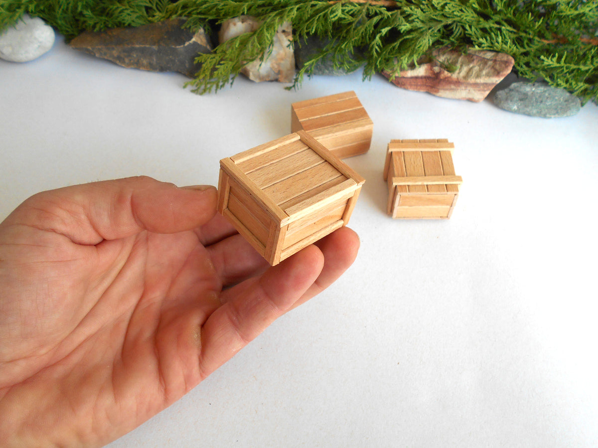 A hand holds a miniature light wood crate, with two smaller matching crates and a larger crate in the background on a white surface with decorative greenery and stones.