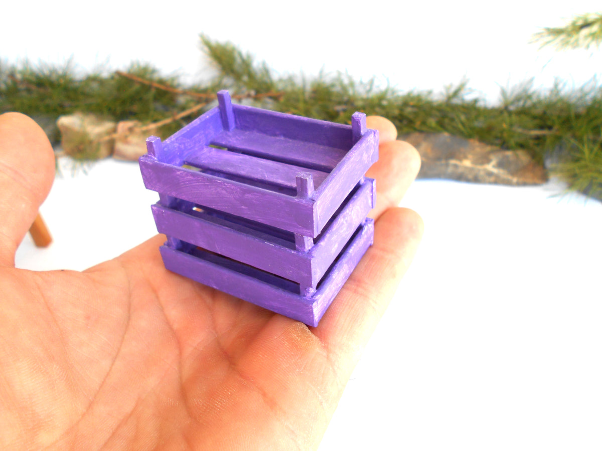 A small purple wooden crate or planter box being held in a person's hand, with a blurred outdoor background of greenery and rocks.