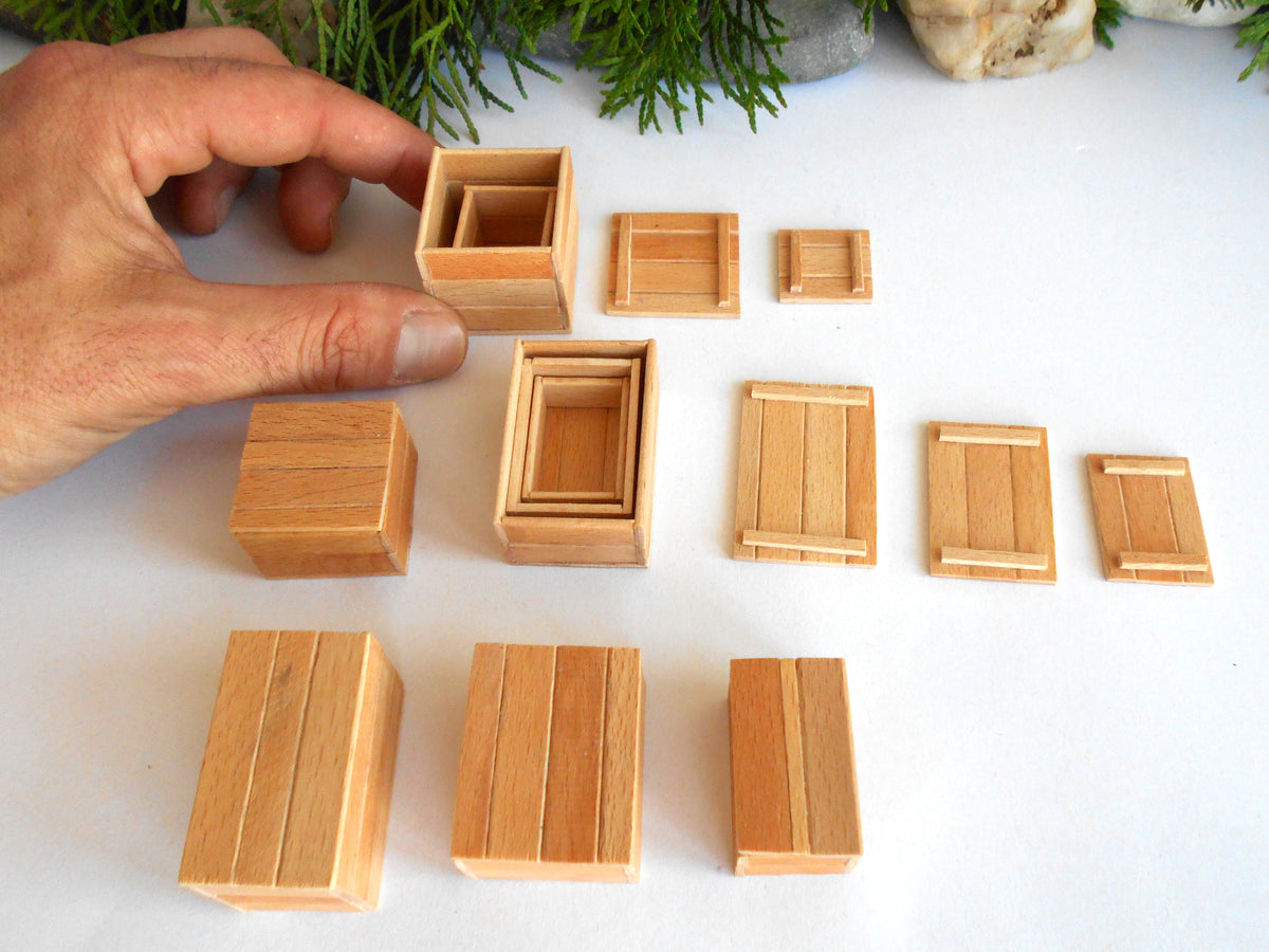A hand is arranging small wooden boxes of different sizes and styles on a white surface, with green foliage and stones in the background.