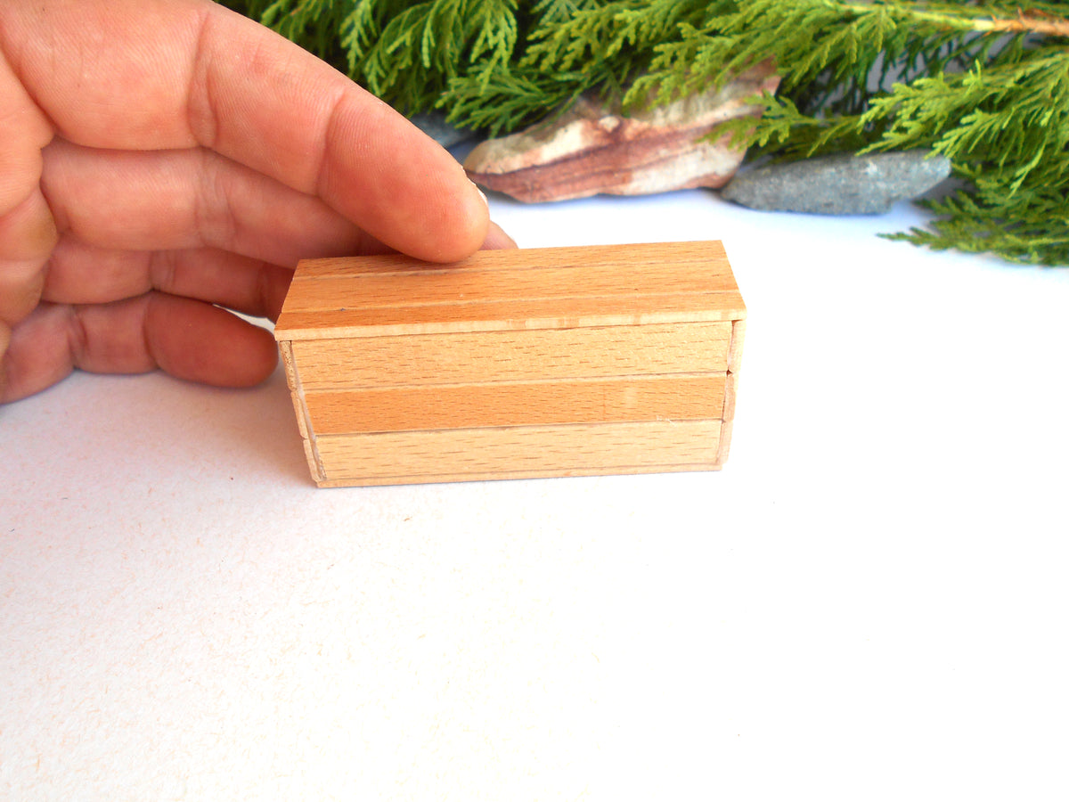 A hand holding a small rectangular wooden box with a striped pattern of light and medium brown wood grain, set against a white background with green foliage and stones in the distance.