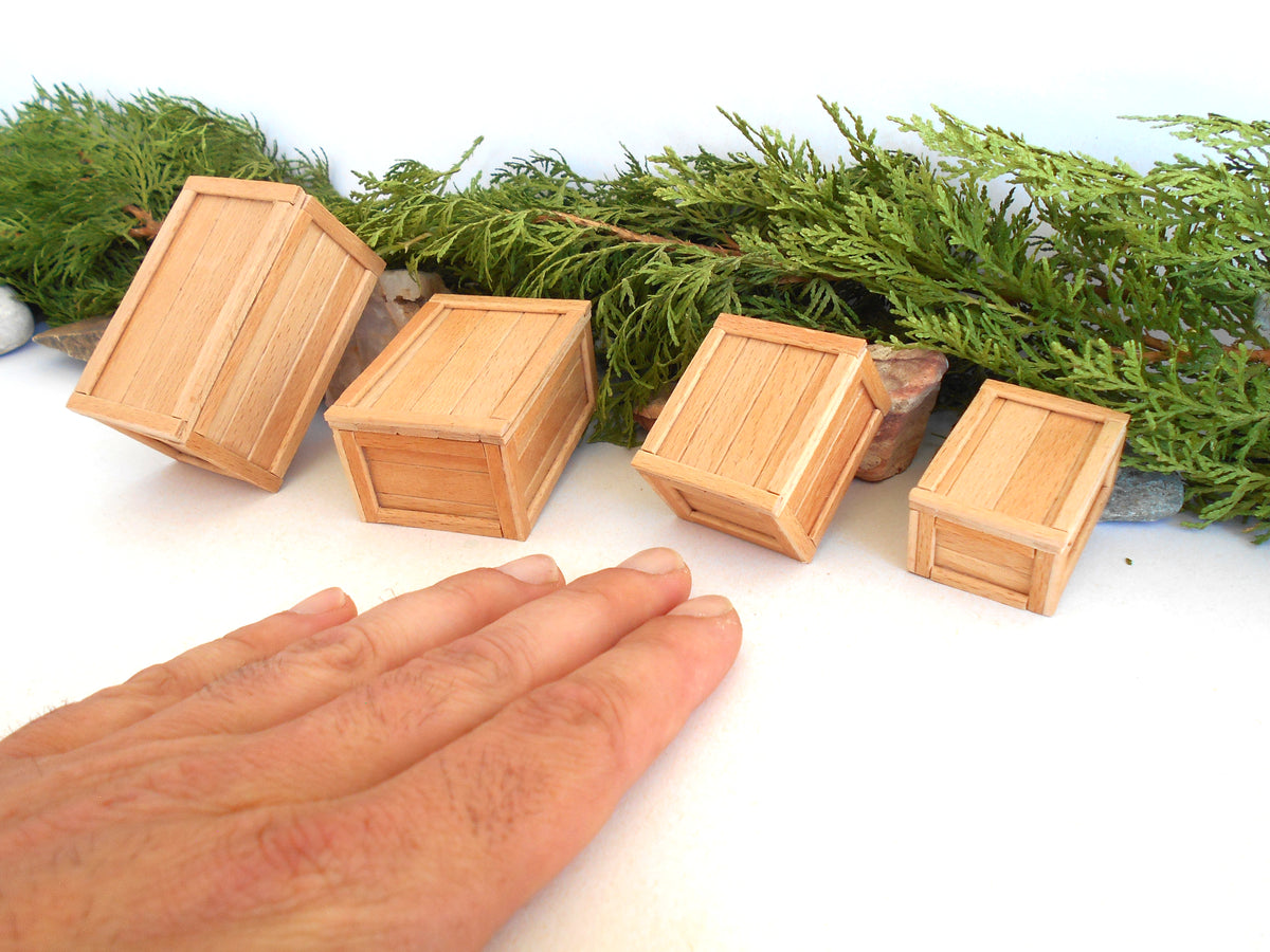 Five miniature light wood boxes of varying sizes arranged on a white surface with green foliage and small stones behind them, and a human hand in the foreground for scale.