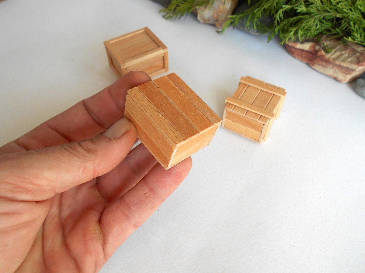 A hand holding a miniature wooden crate, with two more tiny wooden crates in the background on a white surface.