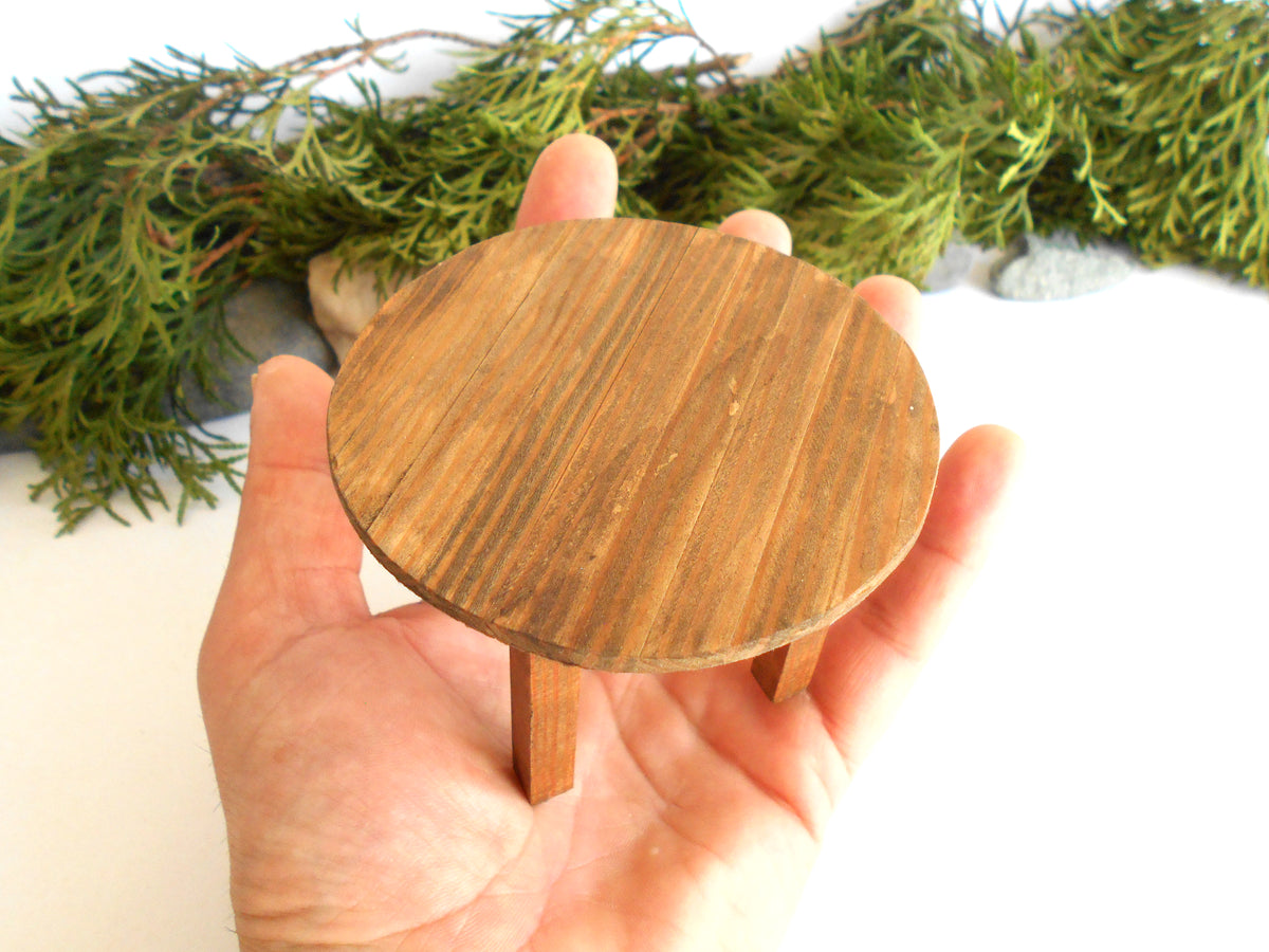 A miniature round wooden table with two legs, held in a person's hand to show its small scale, with green pine branches in the background.
