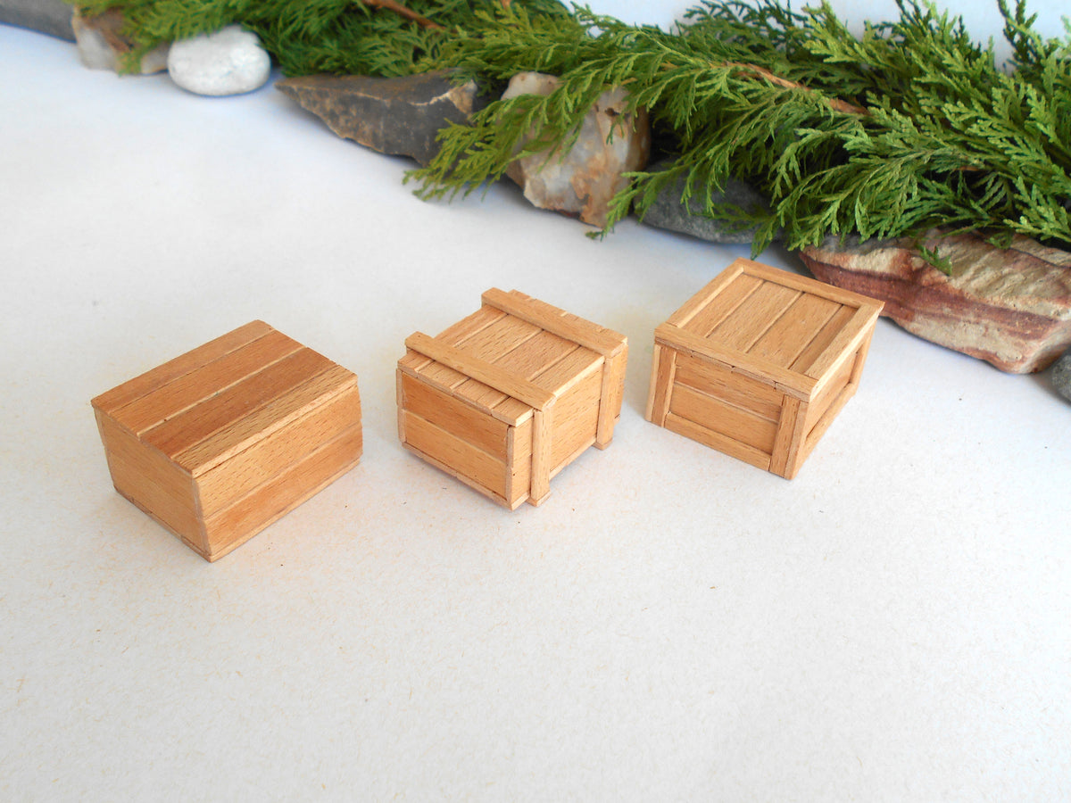 Three miniature light brown wooden crate boxes arranged on a white surface with green foliage and rocks in the background.