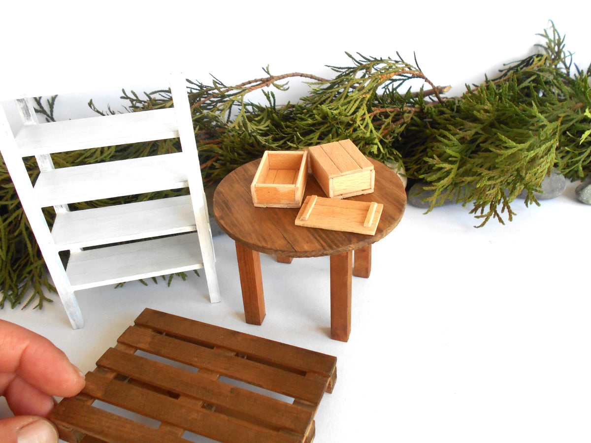 A miniature dollhouse furniture set featuring a round wooden side table with two small wooden boxes, a white wooden chair, and a wooden slatted stool or bench, shown on a white background with green pine branches as styling props. A human hand is holding the bench in the foreground.