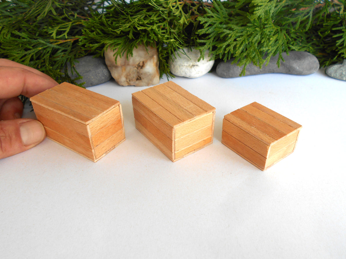 Three small rectangular wooden boxes of different sizes arranged on a white surface, with a hand holding one box on the left and greenery and stones in the background.
