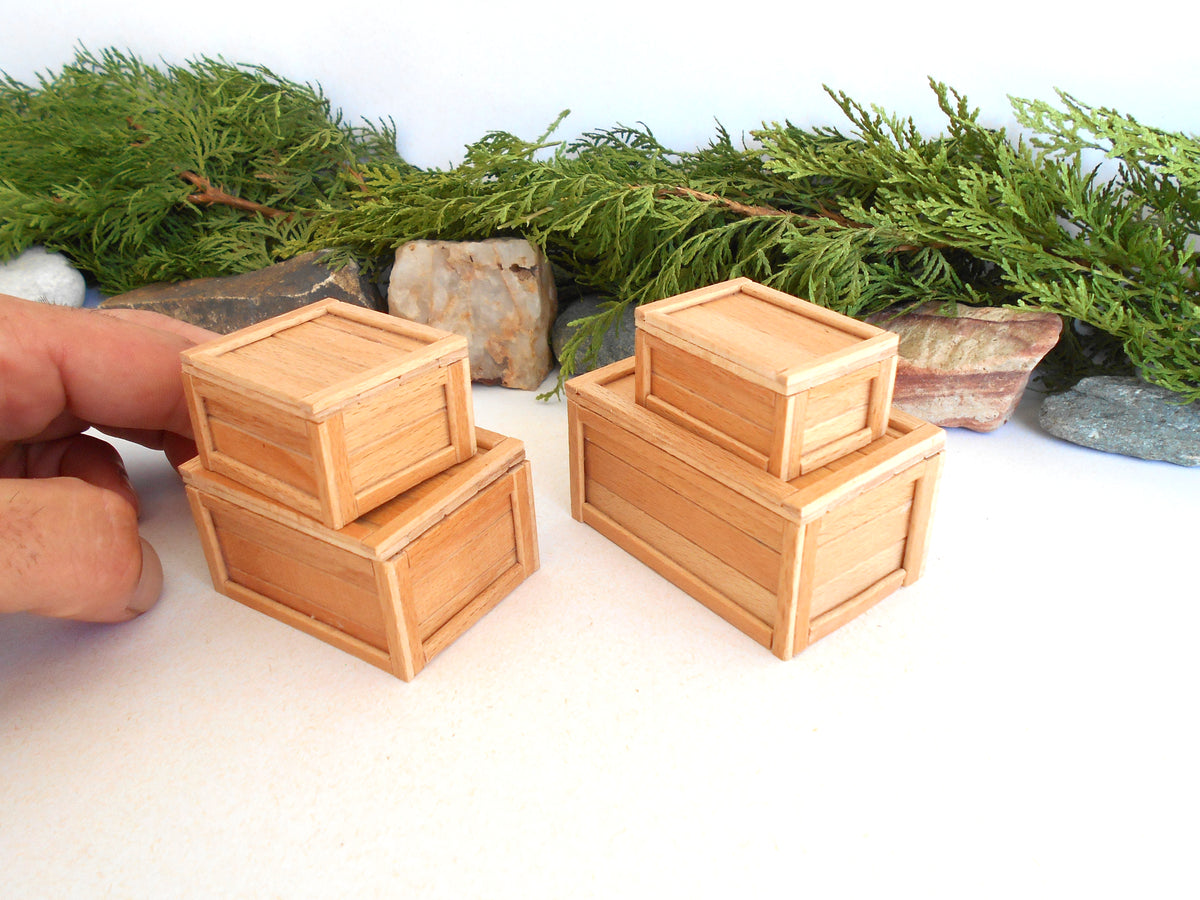 Three miniature light wood crates of different sizes, with a hand holding one of the smaller stacked crates on the left, set against a white background with green foliage and rocks.