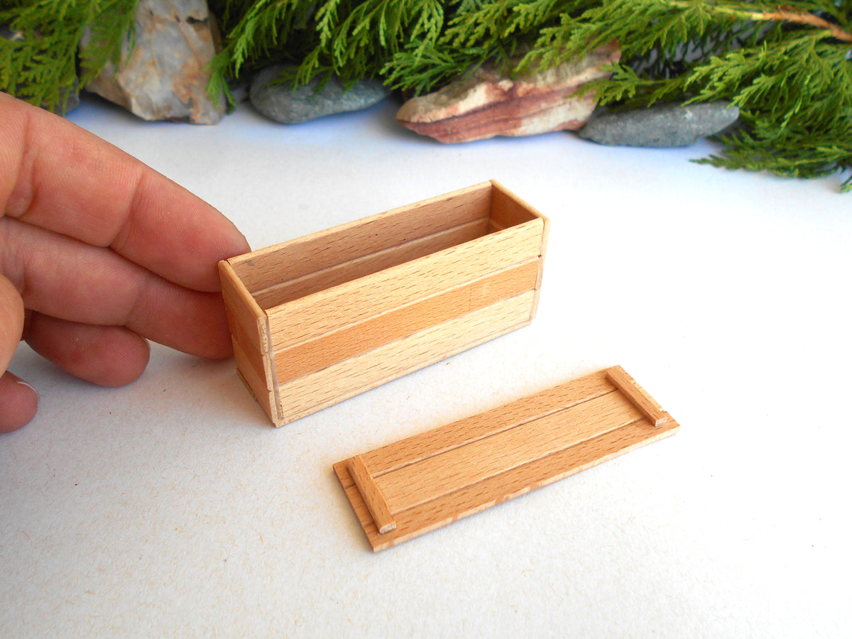 A miniature wooden box with a matching lid, shown on a white surface with a hand holding the box for scale. The background features green foliage and small stones.