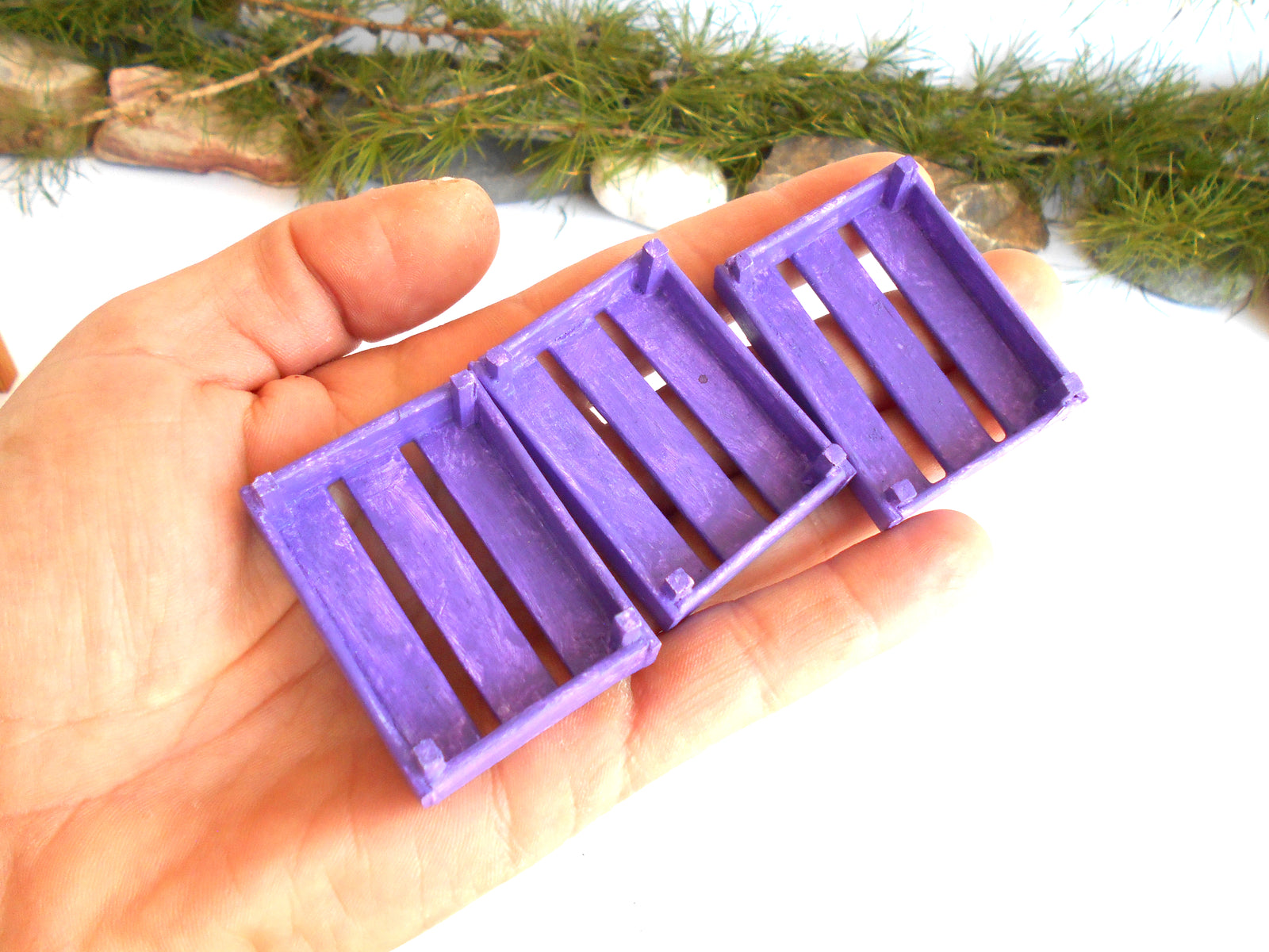 Three small purple plastic crate-style containers held in a person's hand against a white background with blurred greenery at the top.